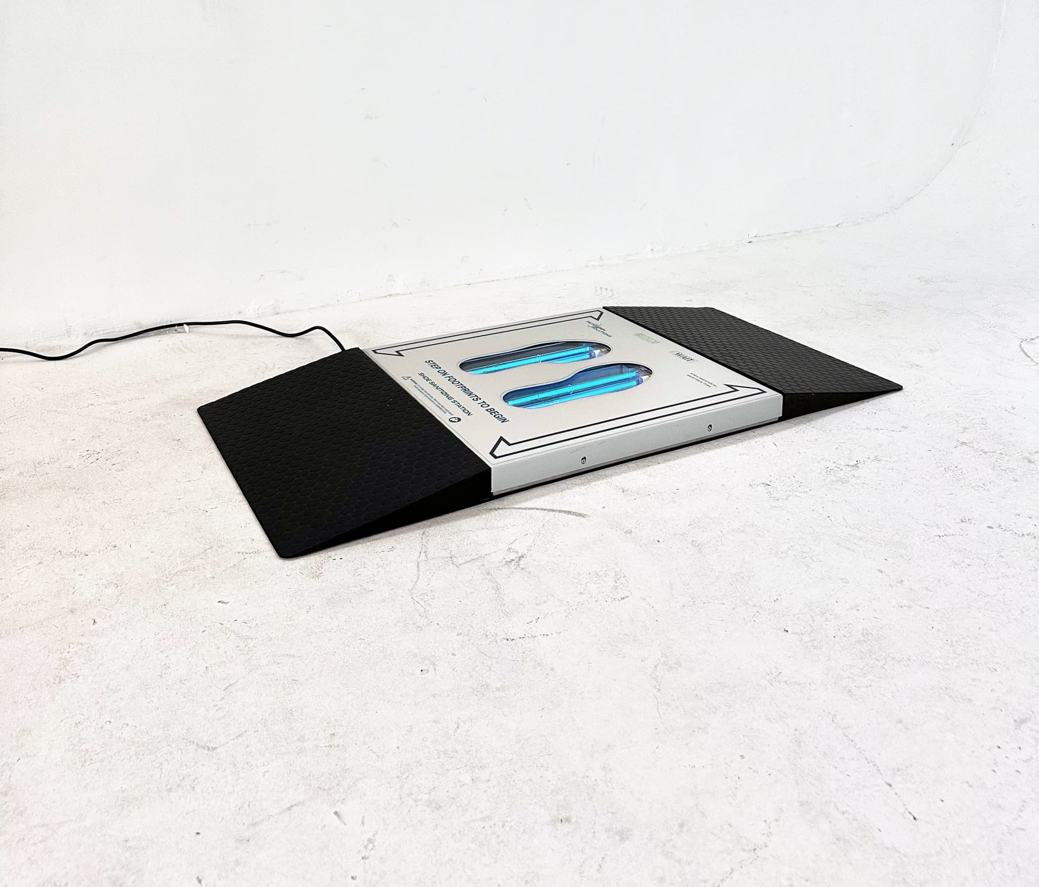 UVZone shoe sanitizing station with a black ramp at each end on a concrete floor, lamps lit behind the two footprint windows, power cord running to the wall