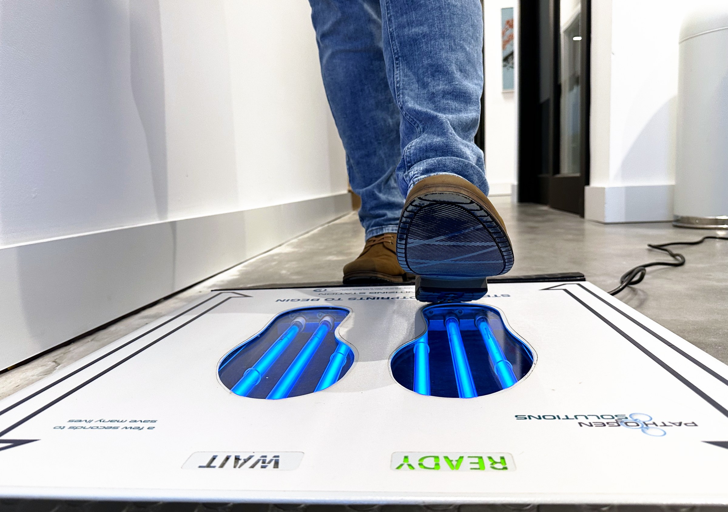 Person in jeans and brown boots stepping across the UVZone shoe sanitizing station, seen from floor level at one end with the READY indicator lit