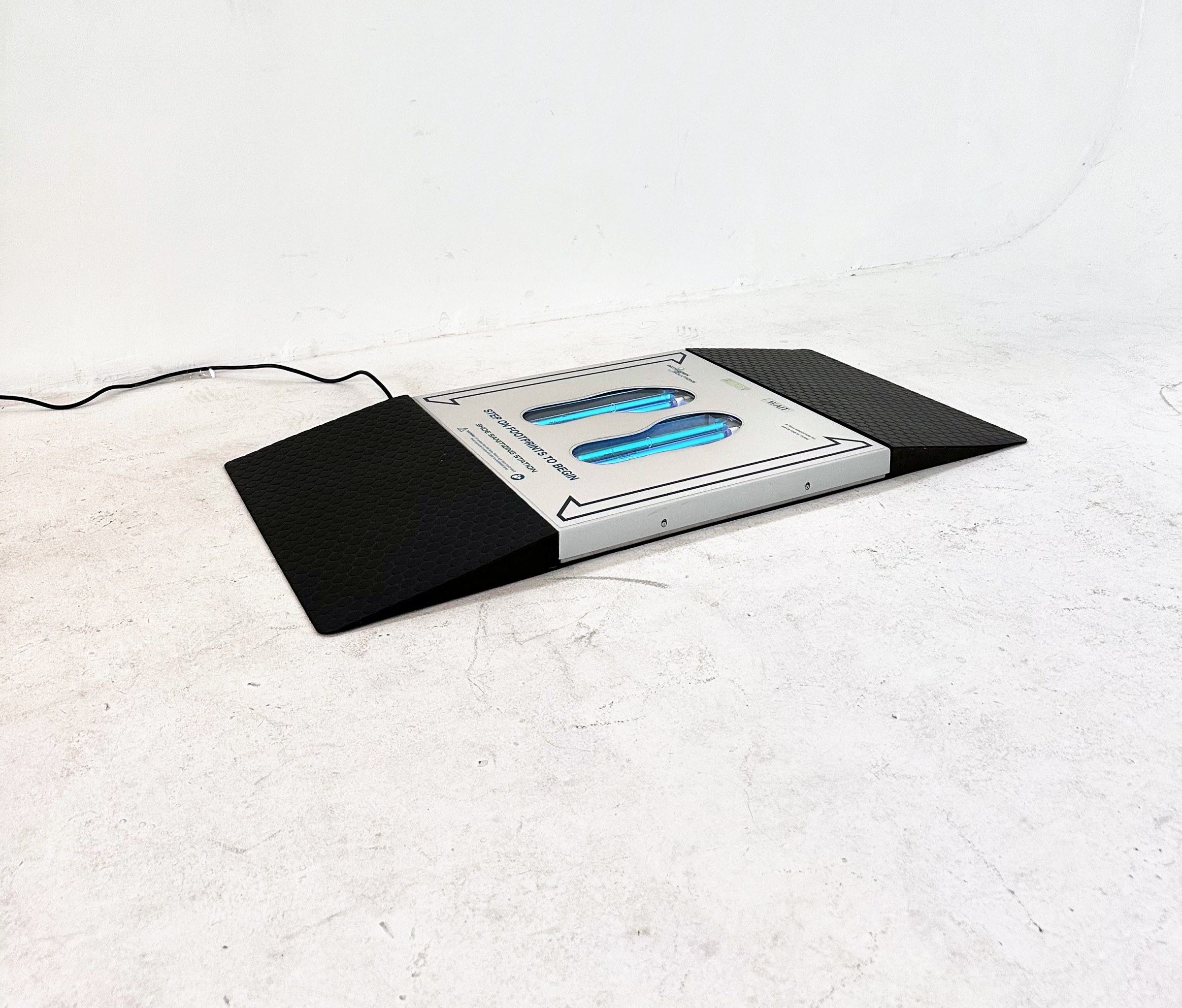 UVZone shoe sanitizing station with a black ramp at each end on a concrete floor, lamps lit behind the two footprint windows, power cord running to the wall