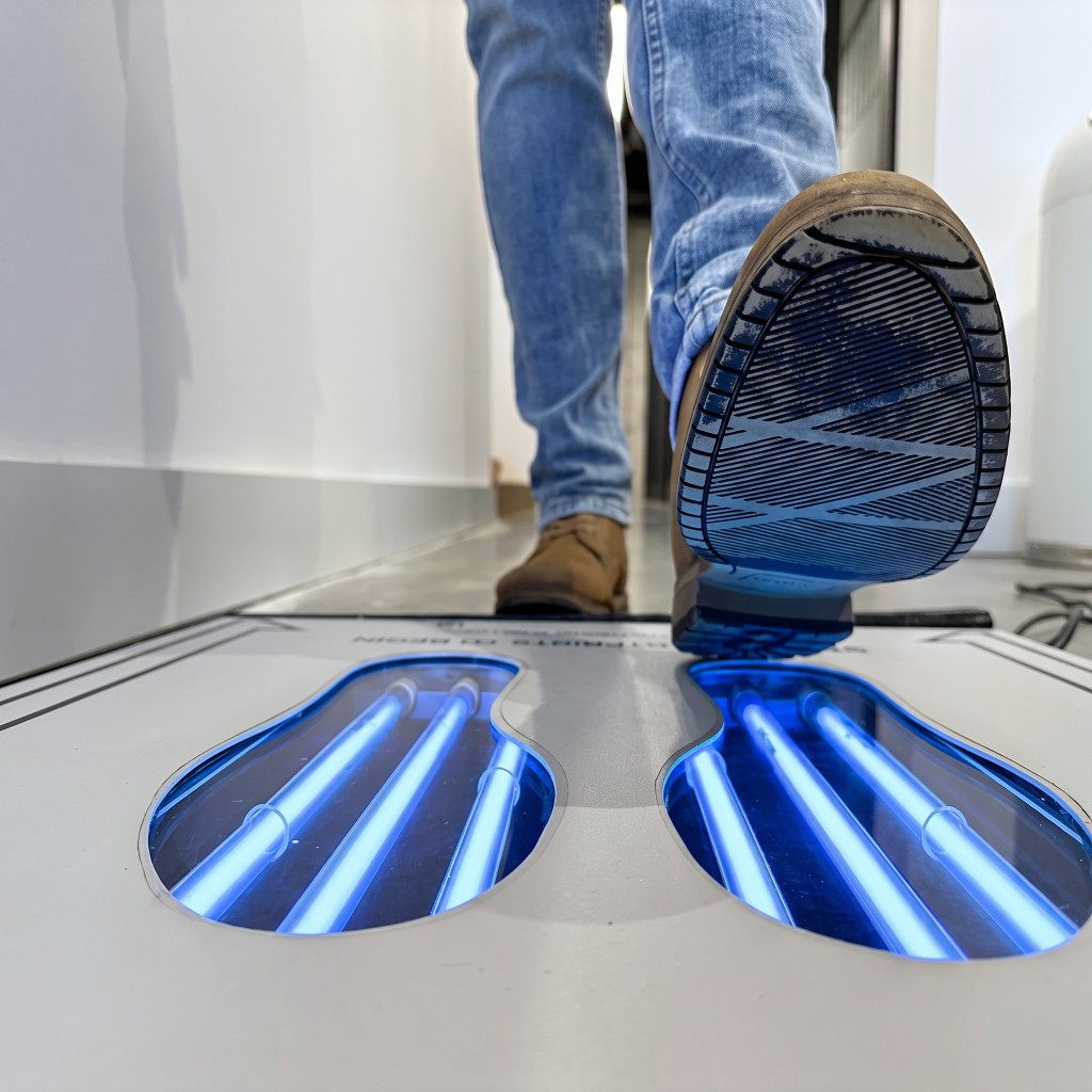 Close-up of a boot sole stepping onto a UVZone shoe sanitizing station with UV-C lights glowing in the footprint cavities